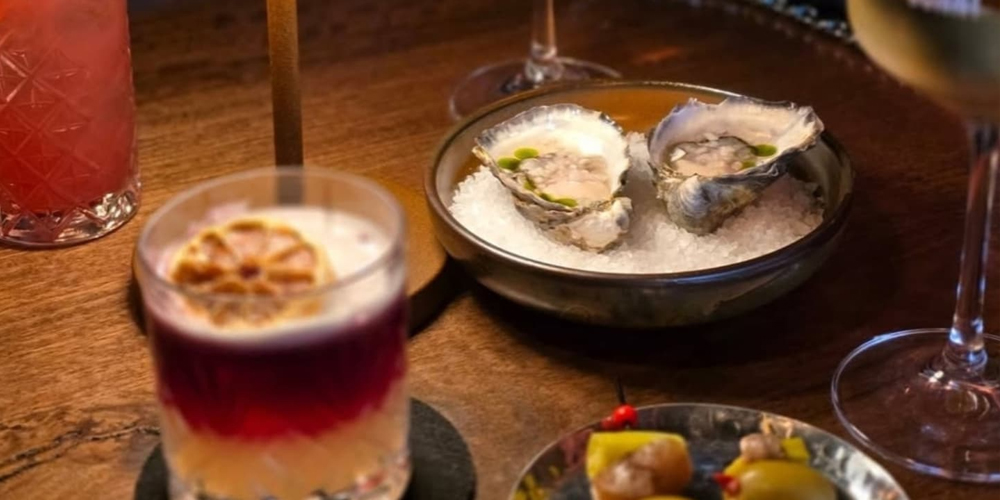 A view of Bonny Bar’s signature bar snacks and cocktails: freshly shucked oysters with yuzu jalapeño dressing, another snack plate, and colourful house cocktails showcasing native Australian ingredients in a warm, moody setting.