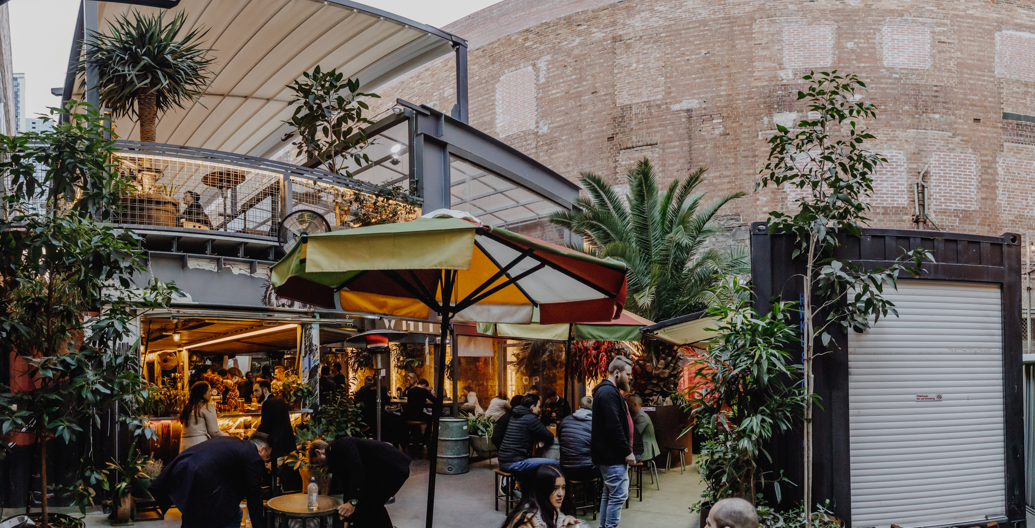 An open-air bar built from stacked shipping containers, adorned with lush greenery and vibrant projections. A lively crowd enjoys craft cocktails, rotating beers, and DJ sets under the city lights.