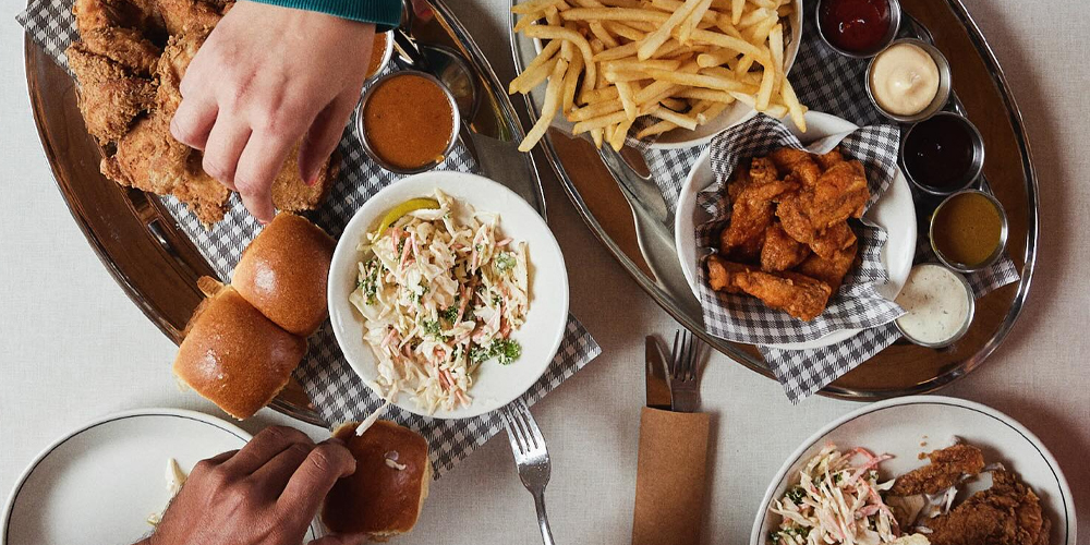 Flatlay of Juanita Peaches’ fried chicken spread featuring golden crispy chicken, buffalo wings, slaw, fries, bread rolls, dipping sauces (honey mustard, boss sauce, ranch, house-made BBQ, and mayo), with diners reaching in.