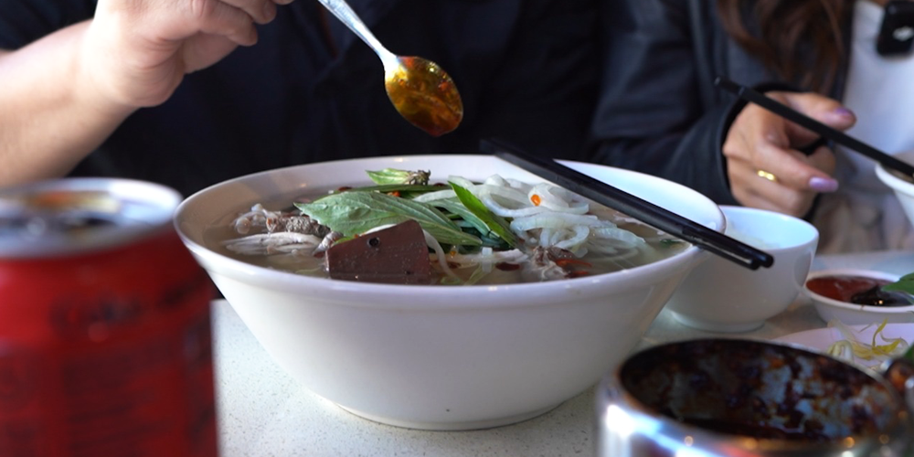 A steaming bowl of traditional Vietnamese pho at Pho Hung Vuong in Footscray, featuring rare beef, fresh herbs, sliced onion, and cubes of beef blood pudding. Diners add chilli oil and condiments for extra flavour. Captured during the morning rush, this hearty noodle soup is a local favourite, served fresh from 9am daily.