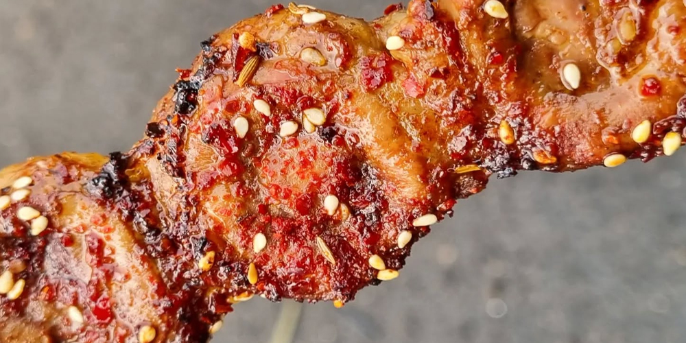 Close-up of grilled lamb skewer at Zi La BBQ in Preston, coated in chilli flakes, cumin seeds, and sesame, charred over charcoal.
