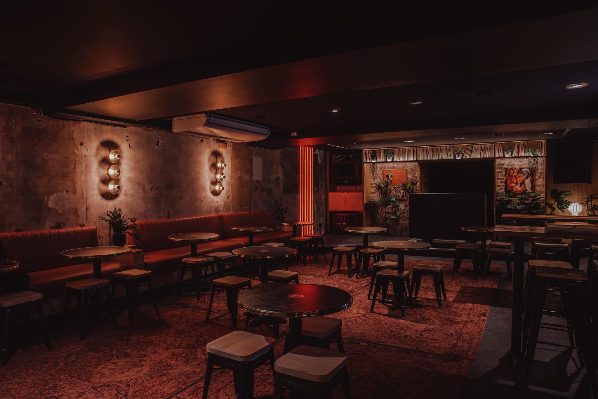 Ambient interior of Wax Music Lounge in Melbourne’s CBD, showcasing its dim red lighting, exposed concrete walls, vintage rugs and intimate tables in a basement on Flinders Street. The photo captures the underground bar’s industrial-chic design, curated lighting, and record-lined stage area that embodies the venue’s Melbourne nightlife and live music culture.